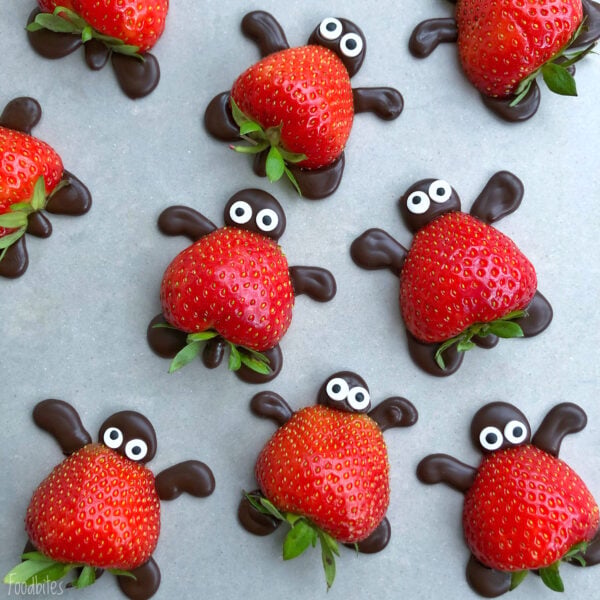 Strawberries decorated as ladybugs with chocolate legs and candy eyes, arranged on a gray surface.