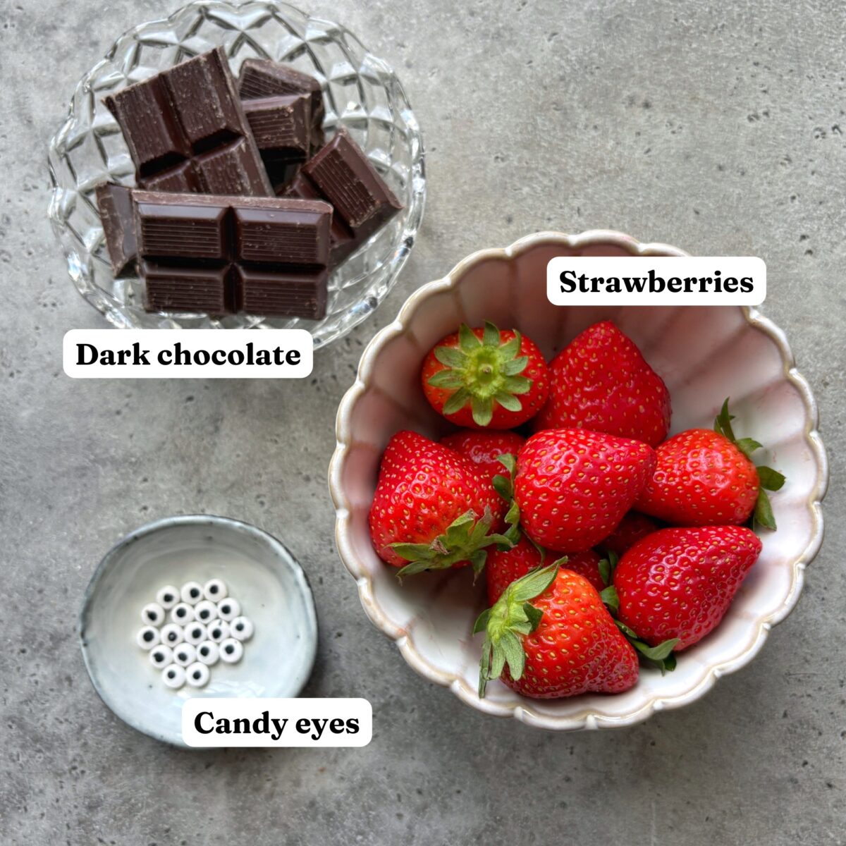 A bowl of strawberries, a dish of dark chocolate squares, and a small dish of candy eyes are arranged on a gray surface.