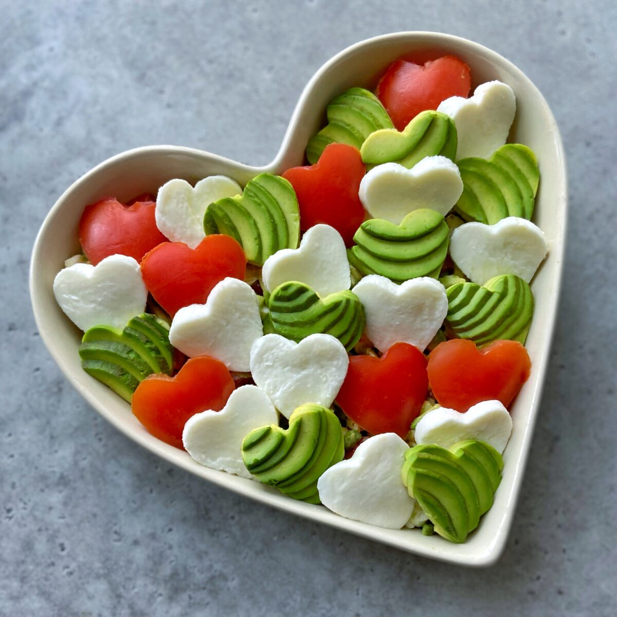 A heart-shaped dish filled with heart-shaped slices of avocado, mozzarella cheese, and tomato, arranged on a gray surface.