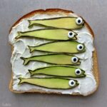 A slice of bread with a layer of cream cheese, topped with six fish-shaped avocado slices.