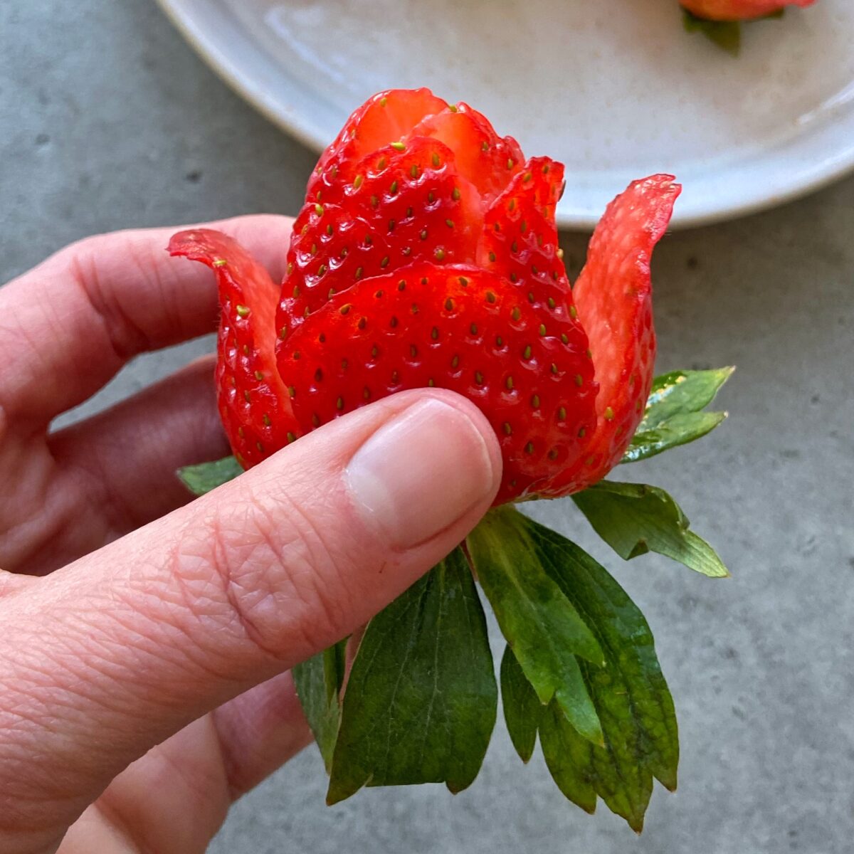 A hand holding a strawberry carved to resemble a rose, with green leaves at the base and a white plate in the background.