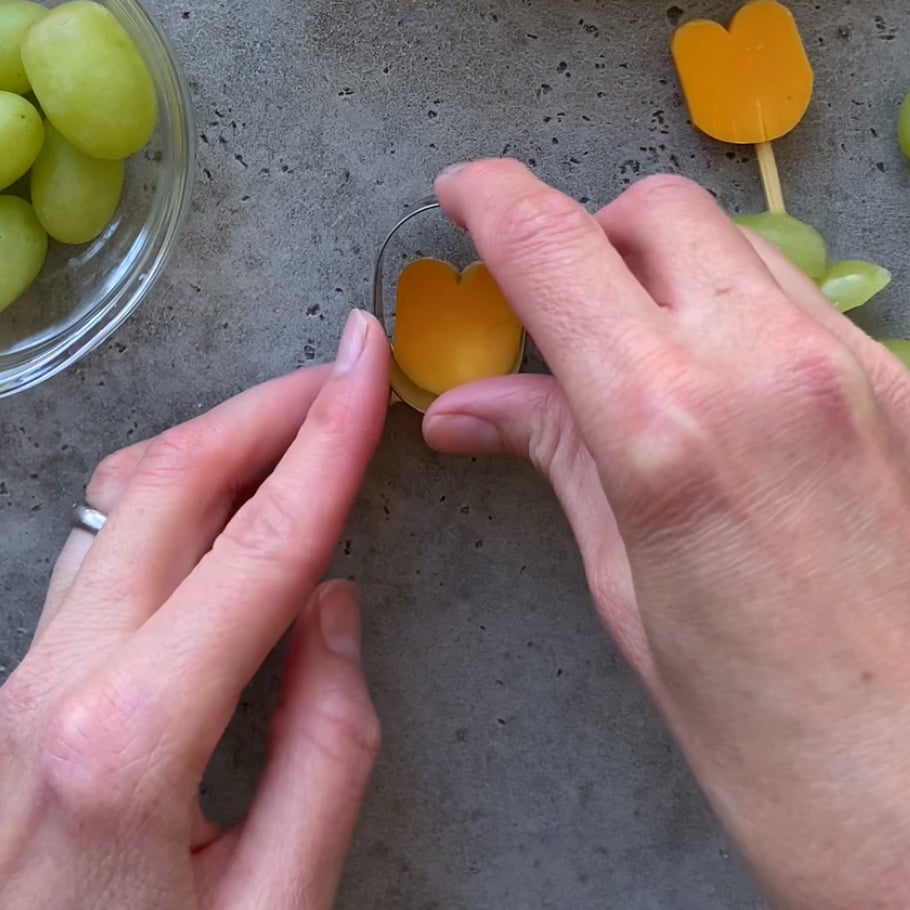 Two hands use a metal cutter to shape a slice of red cheese next to a bowl of green grapes on a gray surface.