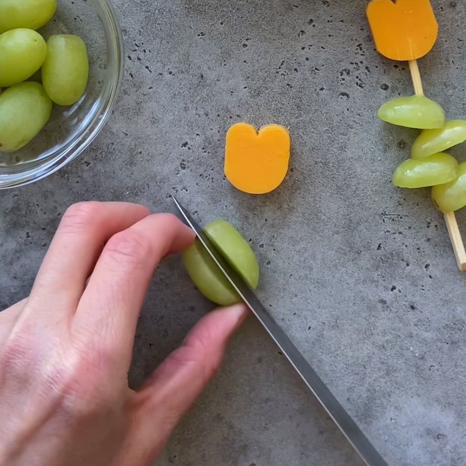 A hand slices a green grape in half with a knife on a gray surface.