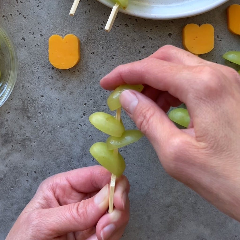 A person prepares Cheese and Grape Skewers by threading green grapes onto a wooden skewer, with tulip-shaped slices of cheddar cheese and a plate visible nearby.