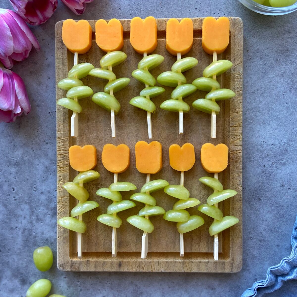 A wooden board displays skewers arranged in two rows, each topped with halved green grapes and a tulip-shaped piece of red cheddar, resembling flowers.