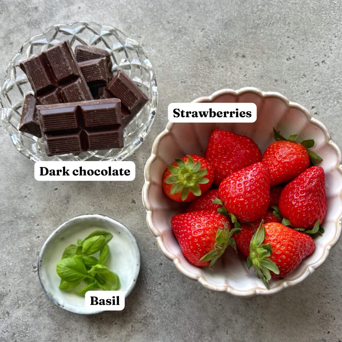 A bowl of strawberries, a dish of dark chocolate pieces, and a small bowl of fresh basil leaves rest on a gray surface. Labels identify each item.