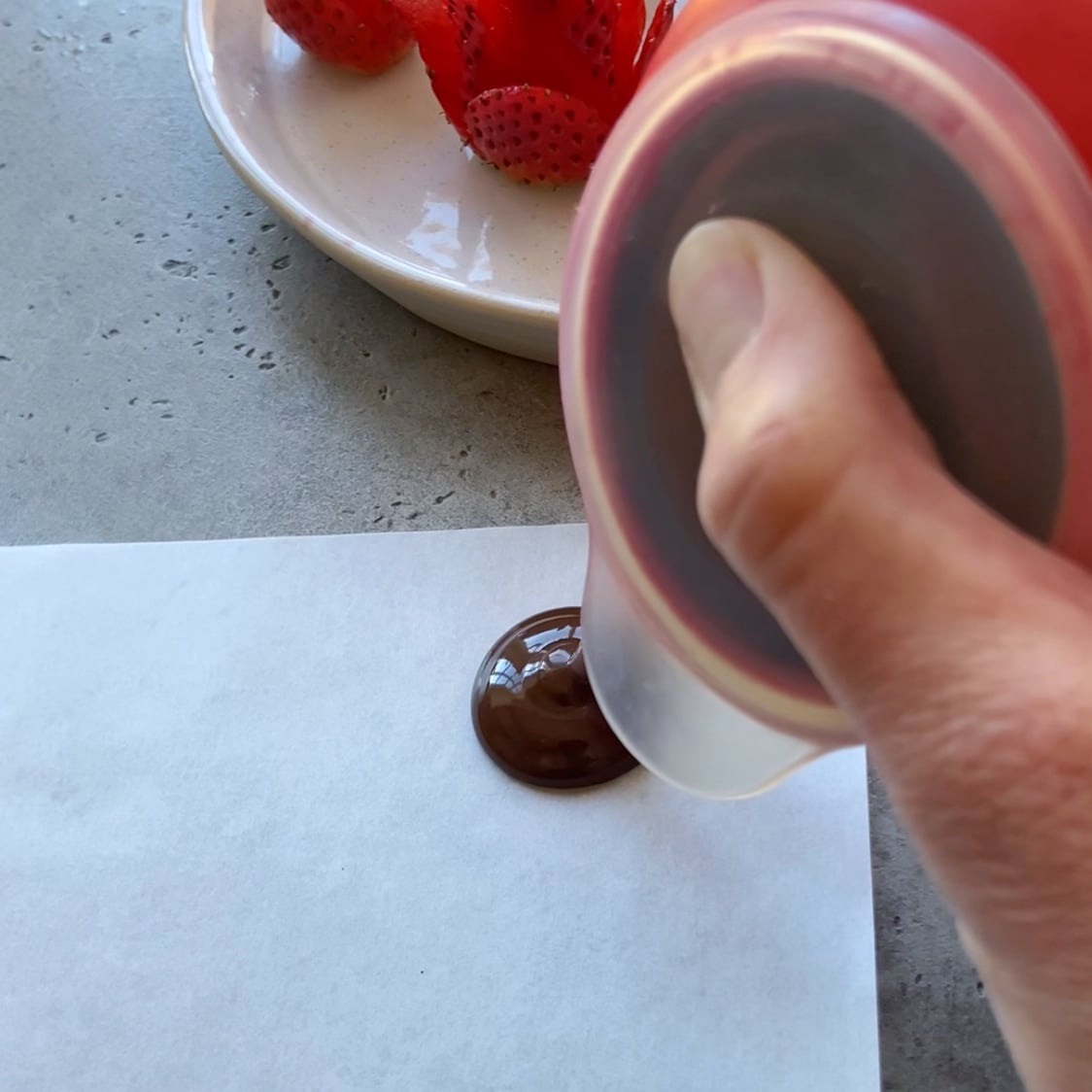 A hand squeezes chocolate onto a piece of white paper, with a plate of Valentine Strawberries sliced in the background.