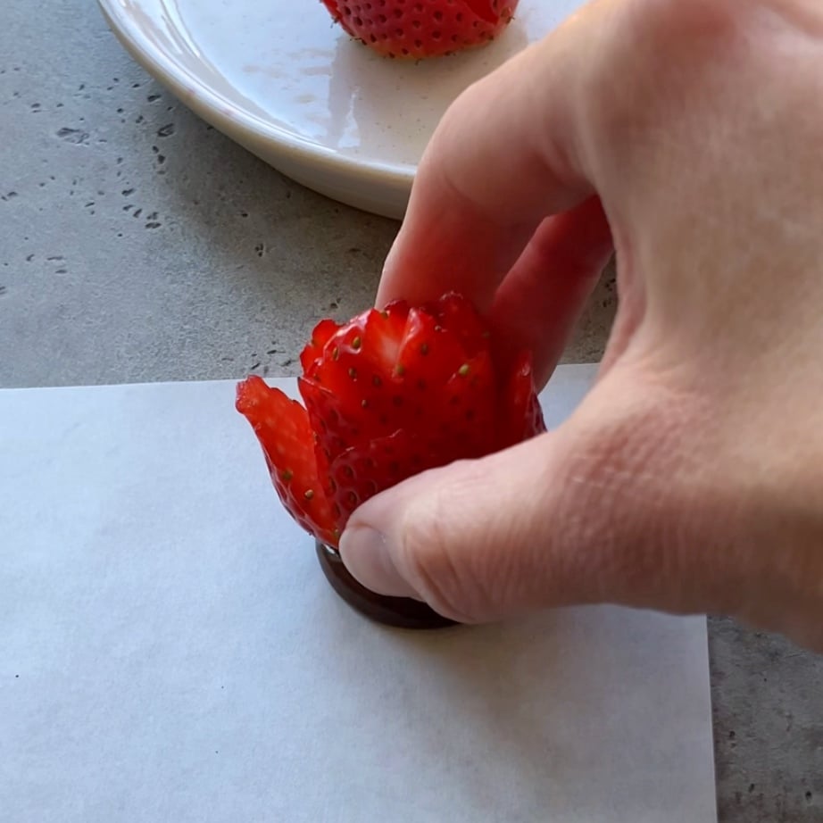A hand arranging a strawberry carved to resemble a flower, placed on a white surface with a plate of Valentine Strawberries in the background.