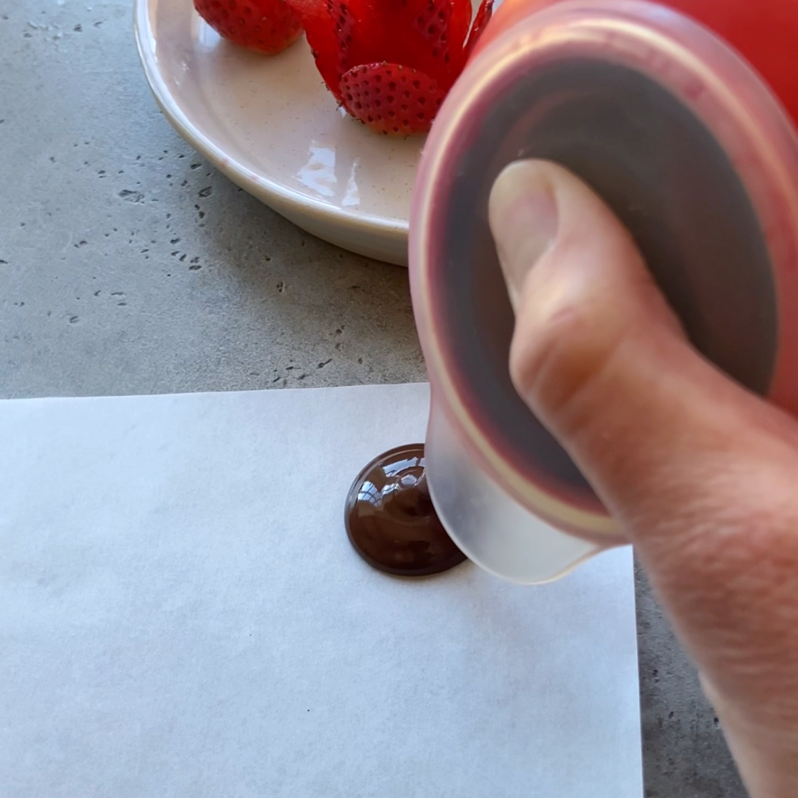 A hand squeezes melted chocolate onto a sheet of white paper, with strawberry roses in the background.