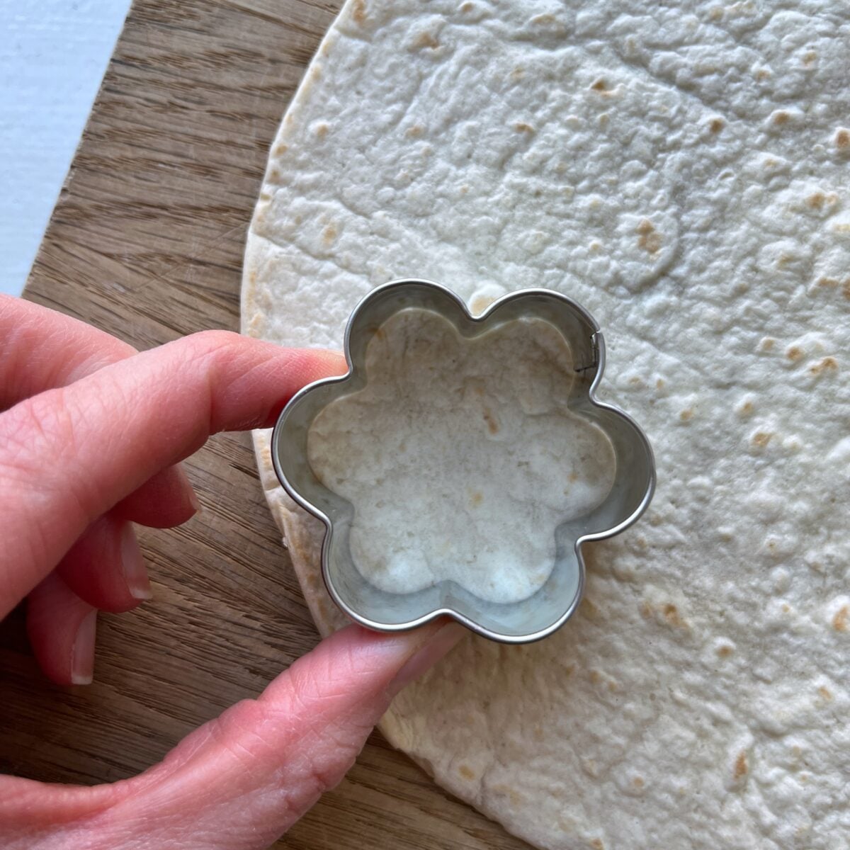 A hand holds a flower-shaped cookie cutter pressing into a tortilla laid on a wooden surface.