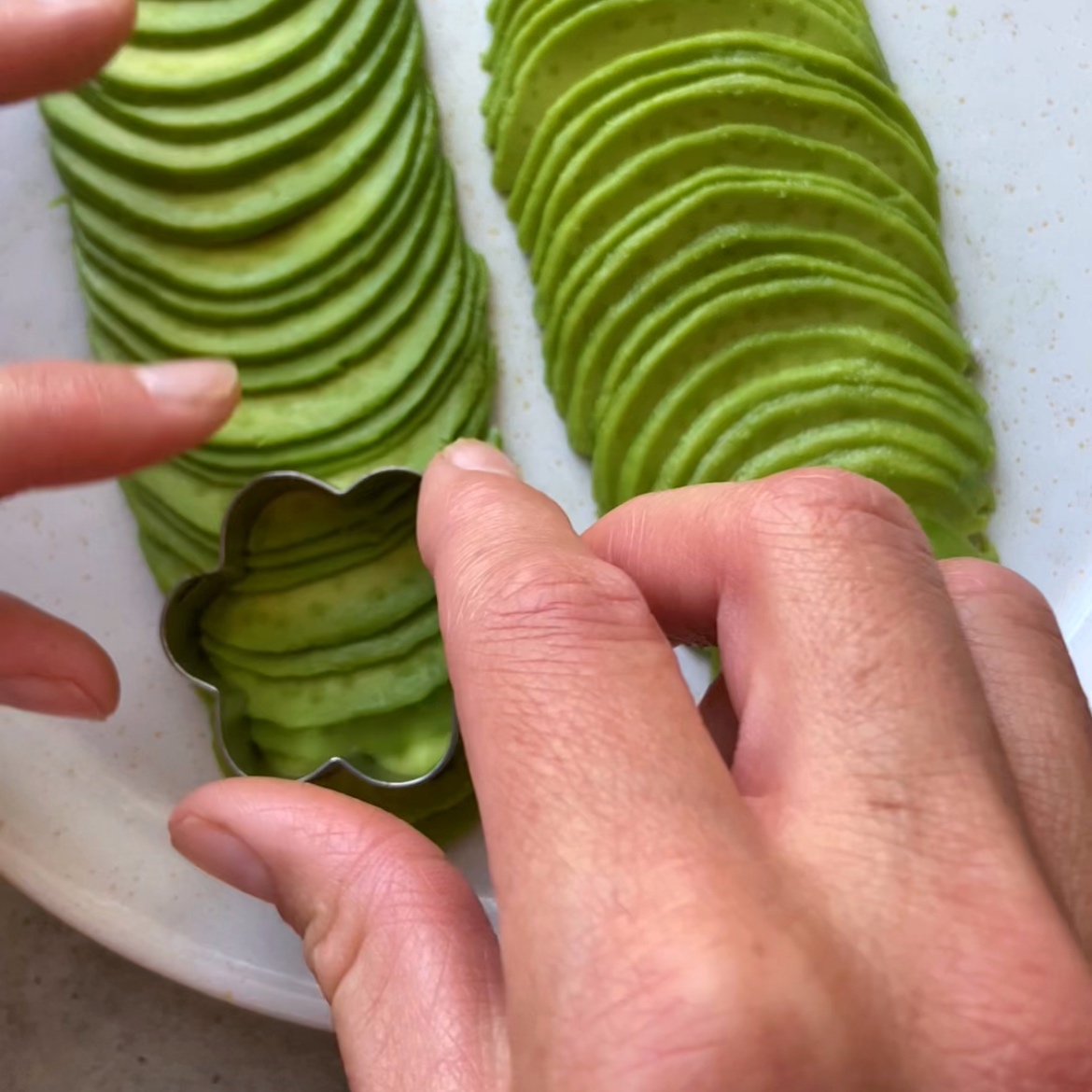A person holds a flower-shaped metal cutter over thinly sliced avocado arranged in neat rows on a plate.