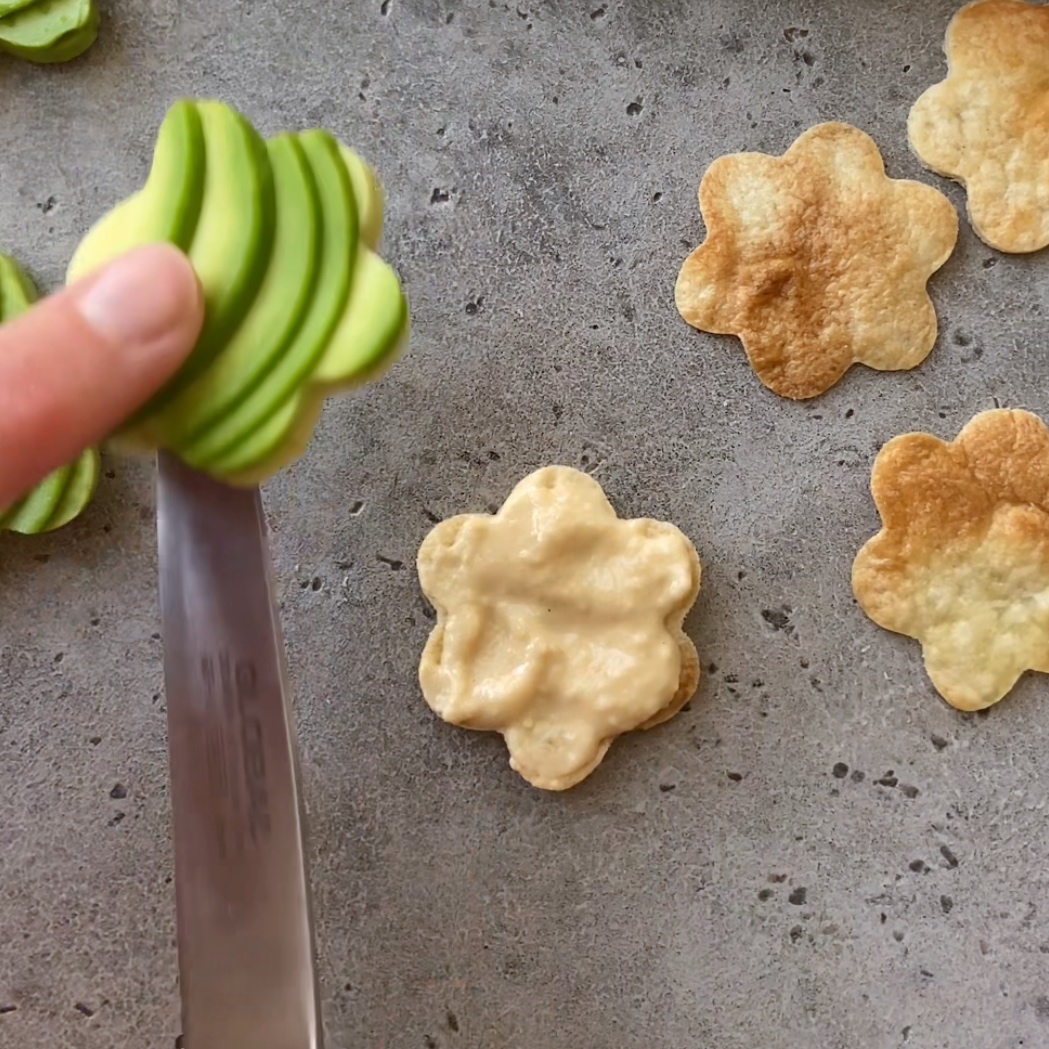 A hand lifts a flower-shaped avocado slice onto a tortilla flower topped with hummus.