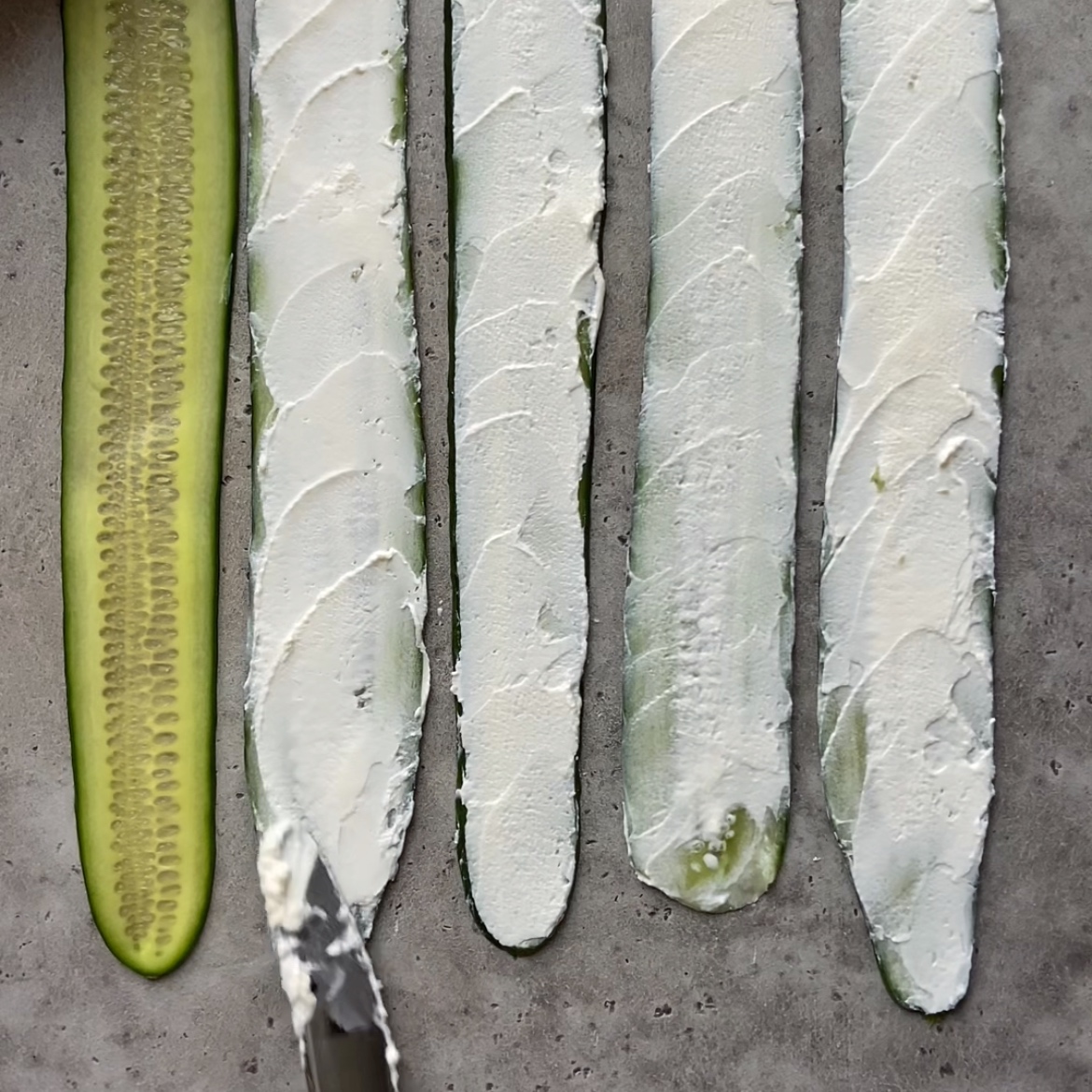 A hand tops cucumber slices with cream cheese.