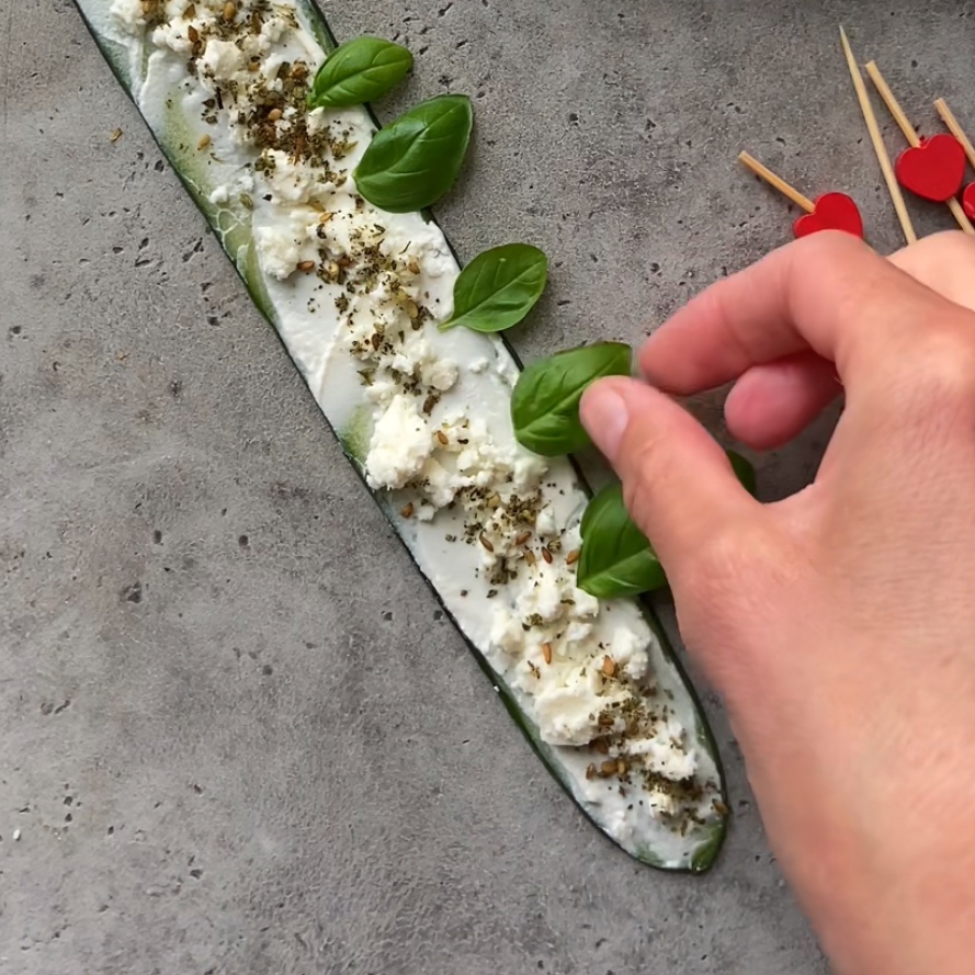 A hand arranges fresh basil leaves onto cucumber strips topped with cream cheese, feta, and zaatar.