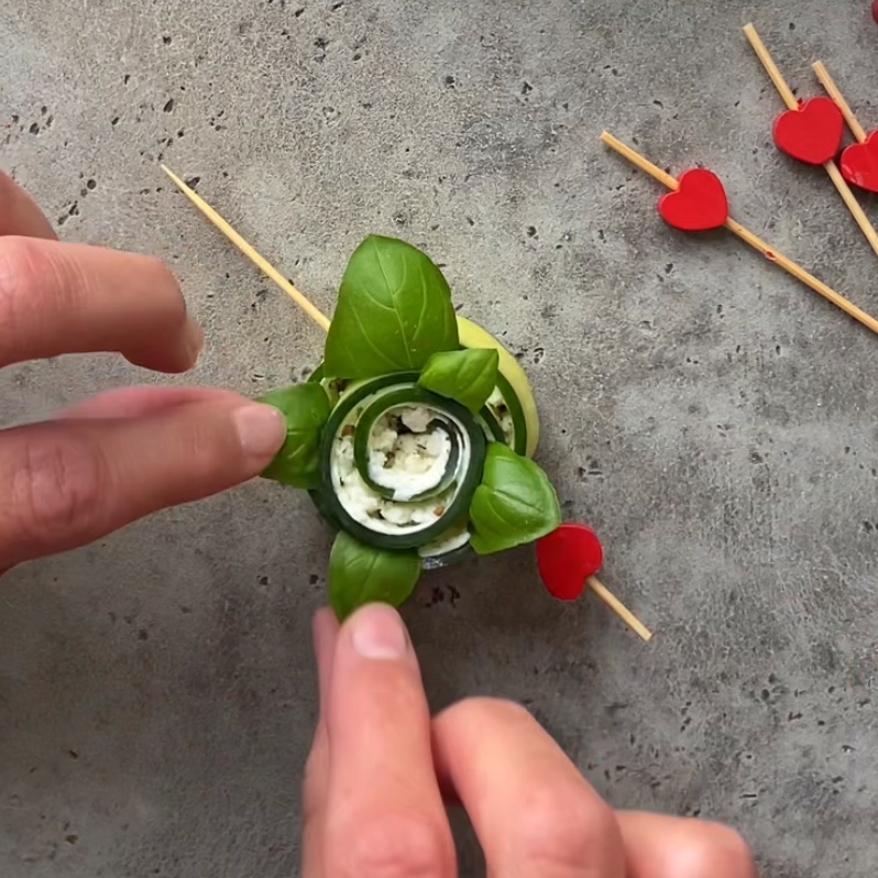 Two hands are folding over fresh basil leaves on a cucumber roll for it to resemble a flower.
