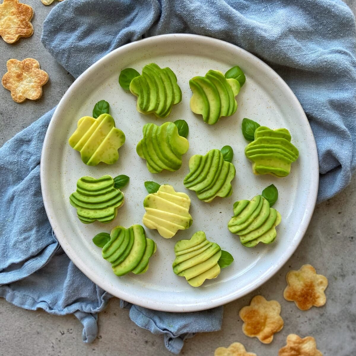 A white plate with eleven fanned avocado bites, decorated with small basil leaves, sits on a blue cloth with flower-shaped tortilla bites scattered nearby.