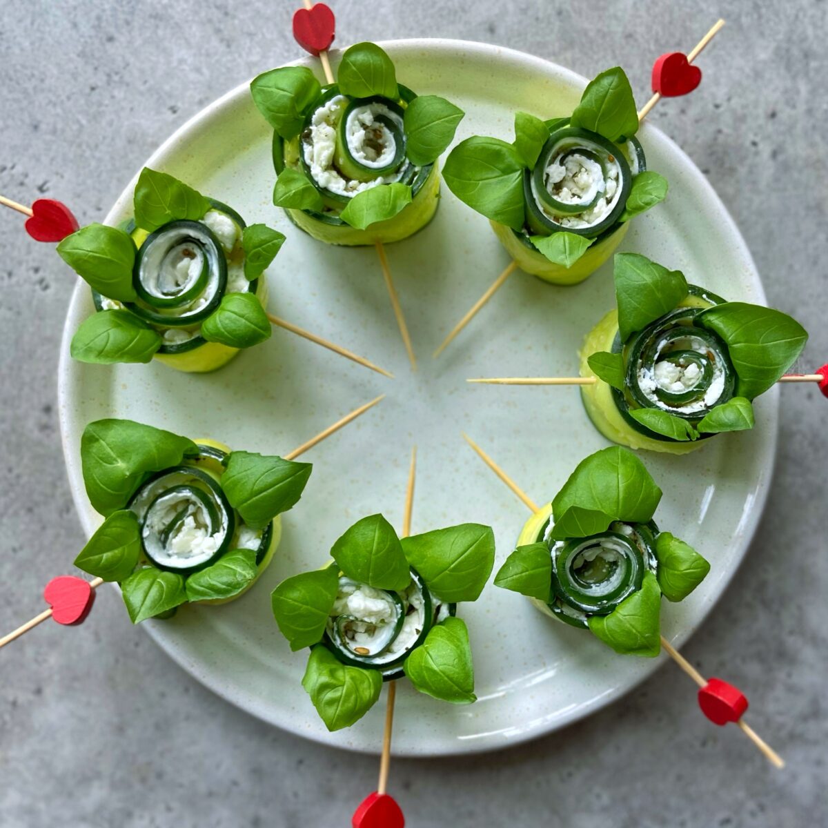 Six cucumber and cheese bites with basil leaves are arranged in a circle on a white plate, each held together with wooden skewers topped with red heart decorations.