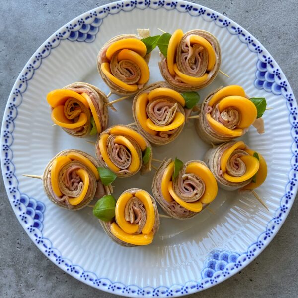 A plate with ten rolled pinwheel bites made of deli meat, cheese, and fresh basil, each held together with a toothpick and arranged in a circular pattern.