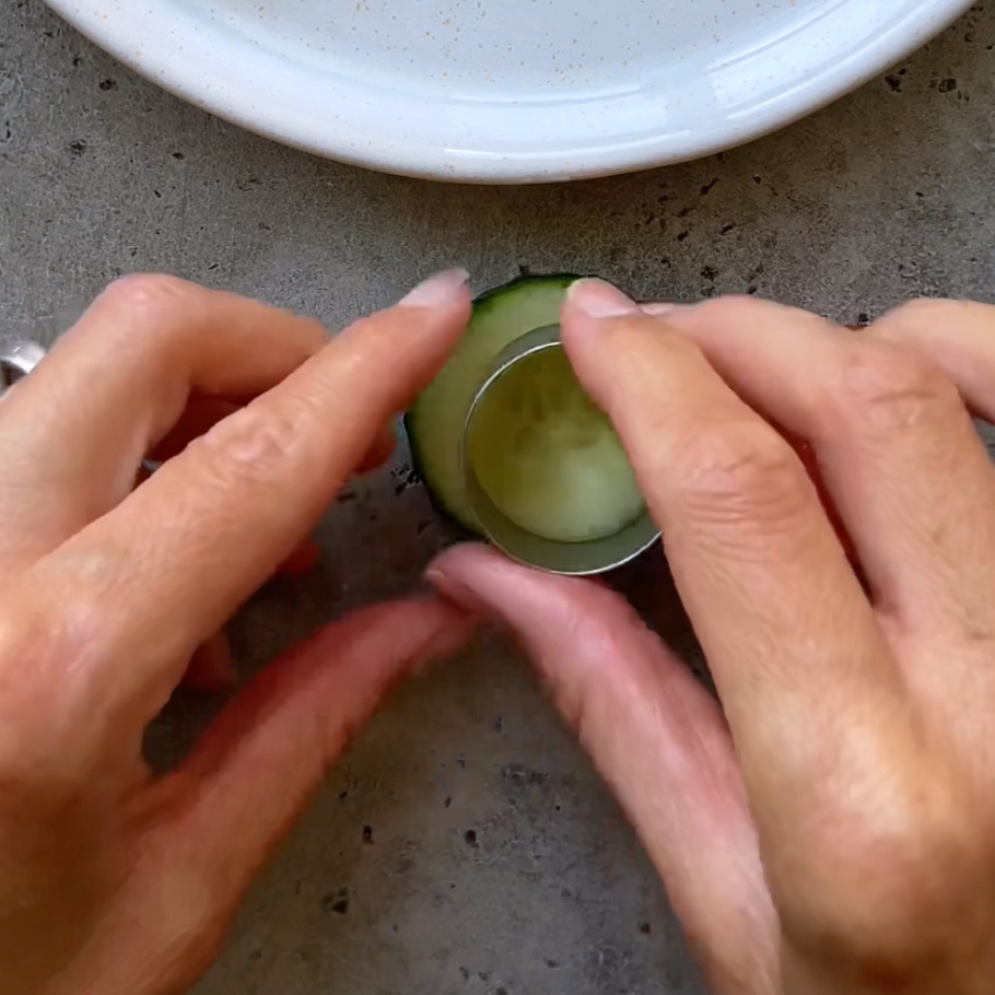Two hands use a round metal cutter to cut out the center of a slice of cucumber.