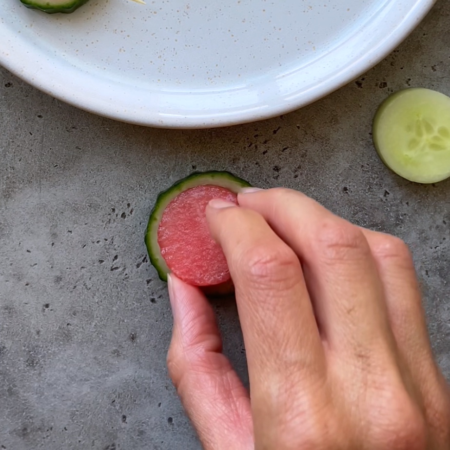 A hand places a thin round slice of watermelon inside a cucumber slice.
