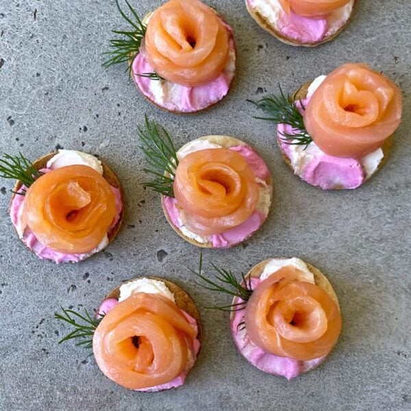 Smoked Salmon Canapés featuring round tortilla crackers topped with pink and white cream cheese, and smoked salmon shaped like roses, garnished with fresh dill on a gray surface.