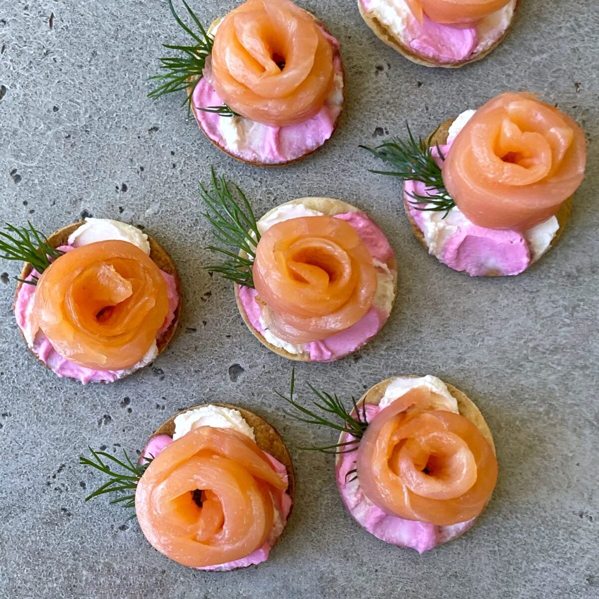 Round tortillas topped with pink and white cream cheese, salmon rosettes, and small sprigs of dill.