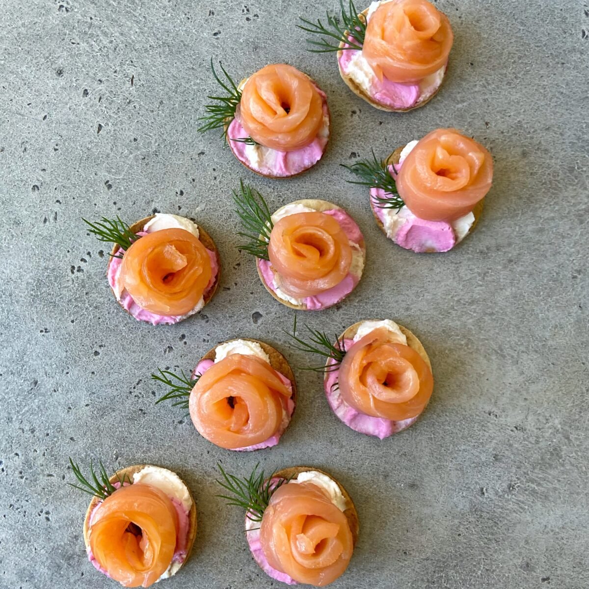 Nine smoked salmon canapés feature tortilla discs topped with pink and white cream cheese, smoked salmon arranged in a rose shape, and garnished with dill.