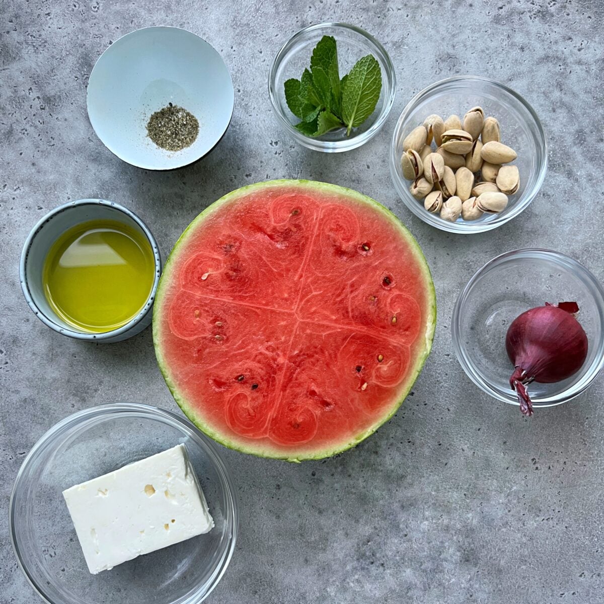 A halved watermelon surrounded by bowls of feta cheese, olive oil, black pepper, mint leaves, pistachios, and a small red onion on a grey surface.