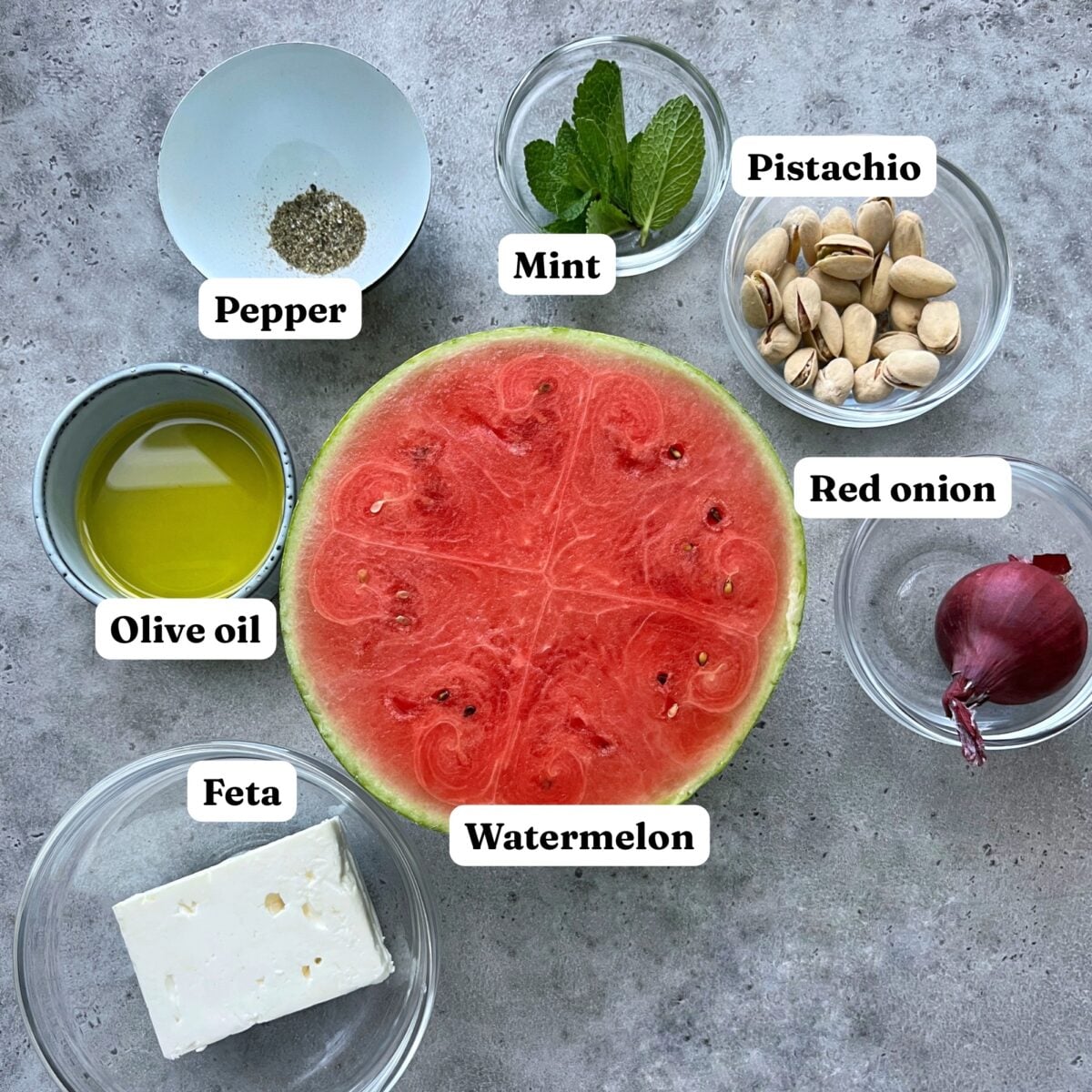A halved watermelon surrounded by bowls of feta cheese, olive oil, black pepper, mint leaves, pistachios, and a small red onion on a grey surface.