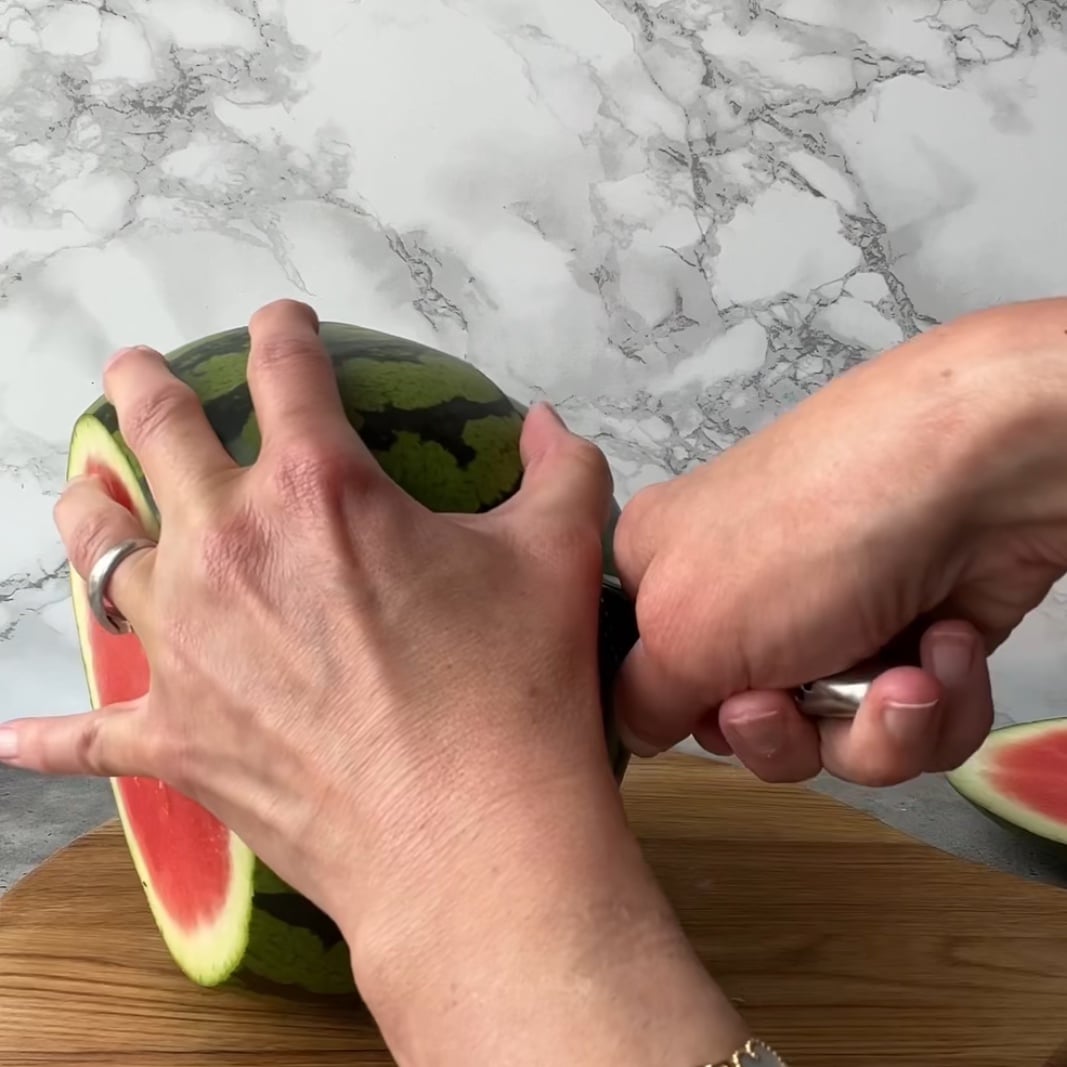 A person slices the sides off a watermelon with a knife on a wooden surface.