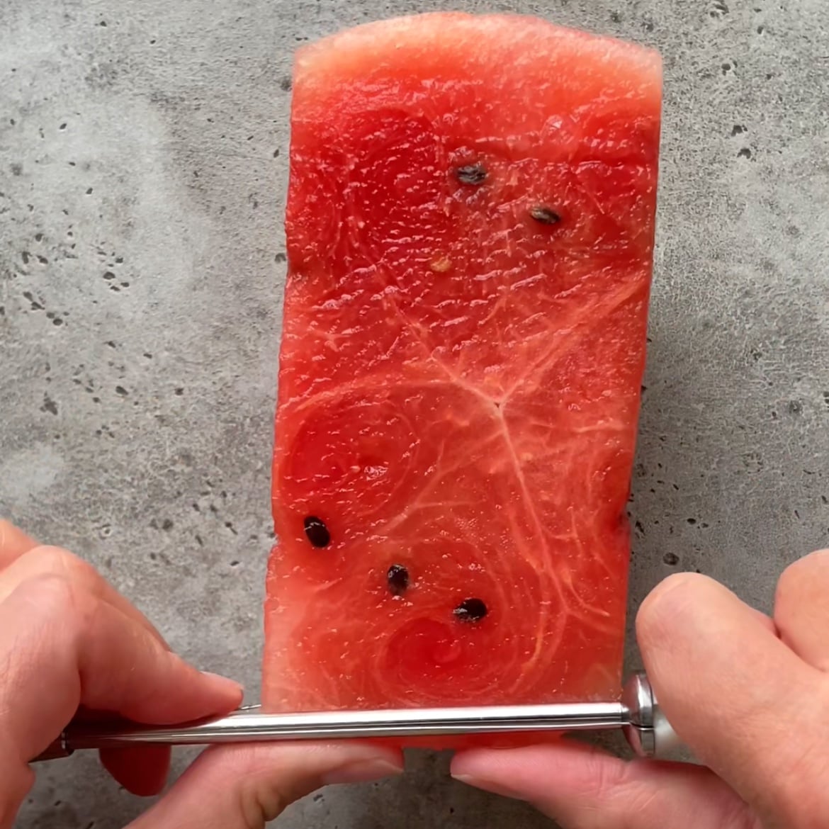 A person slices a thin piece of watermelon on a gray surface.
