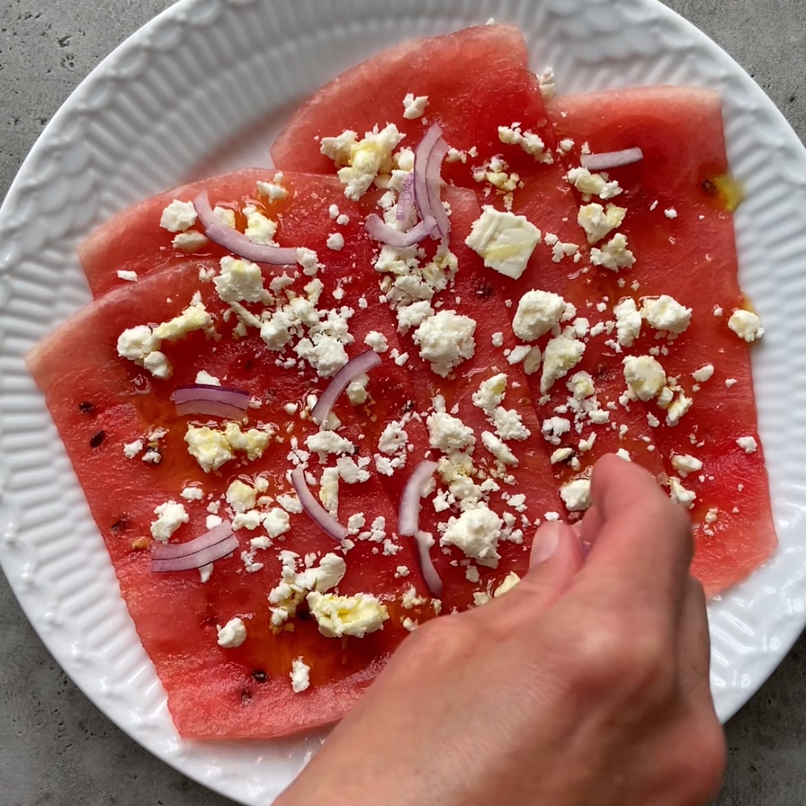 A hand sprinkles crumbled feta cheese over thin slices of watermelon and red onion.