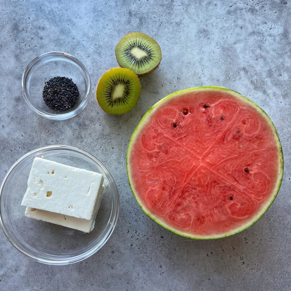 A halved watermelon, two kiwi halves, a bowl of feta cheese, and a bowl of black sesame seeds on a gray surface.