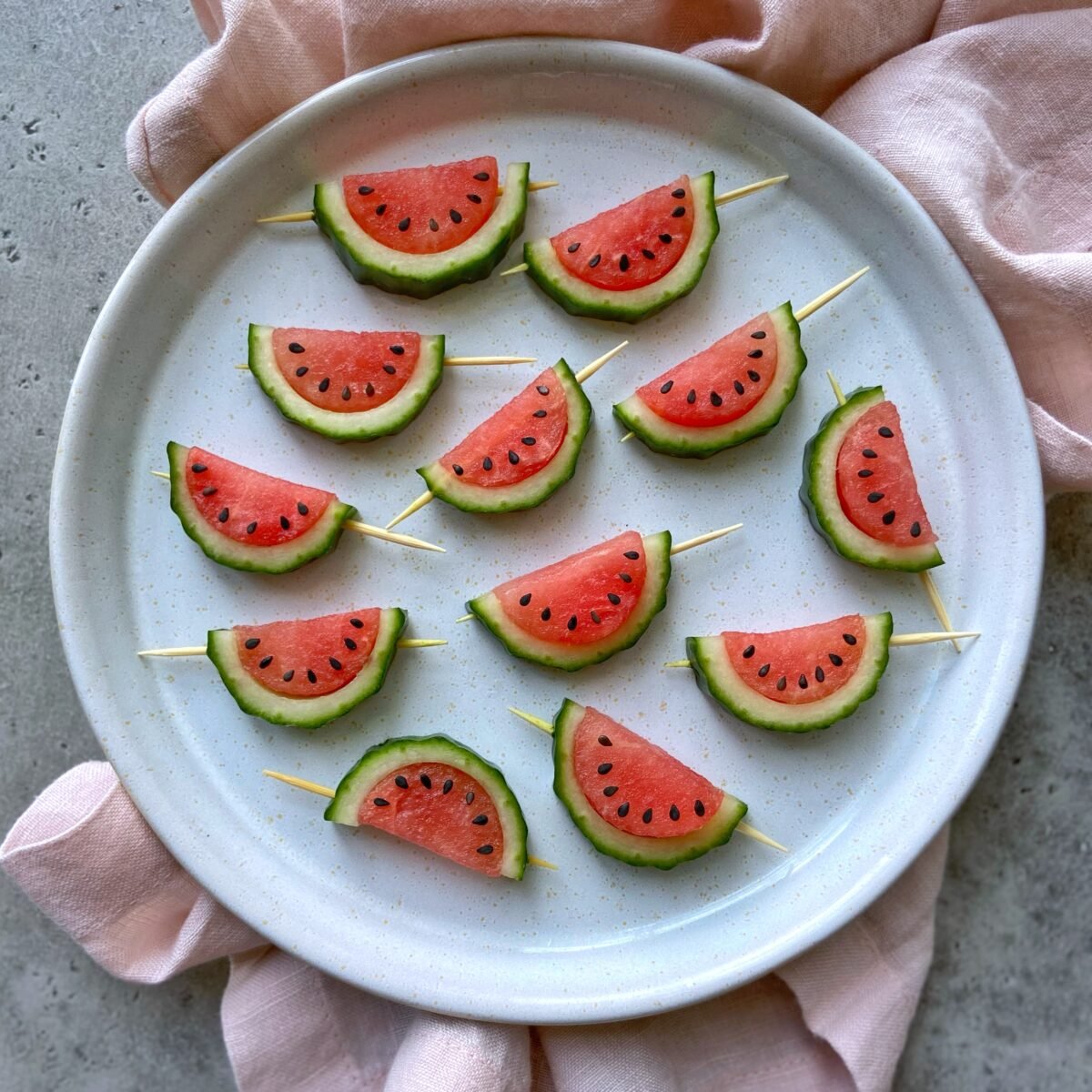 A round plate featuring cucumber and watermelon on skewers, arranged to resemble mini watermelon slices, sits on a light pink cloth.
