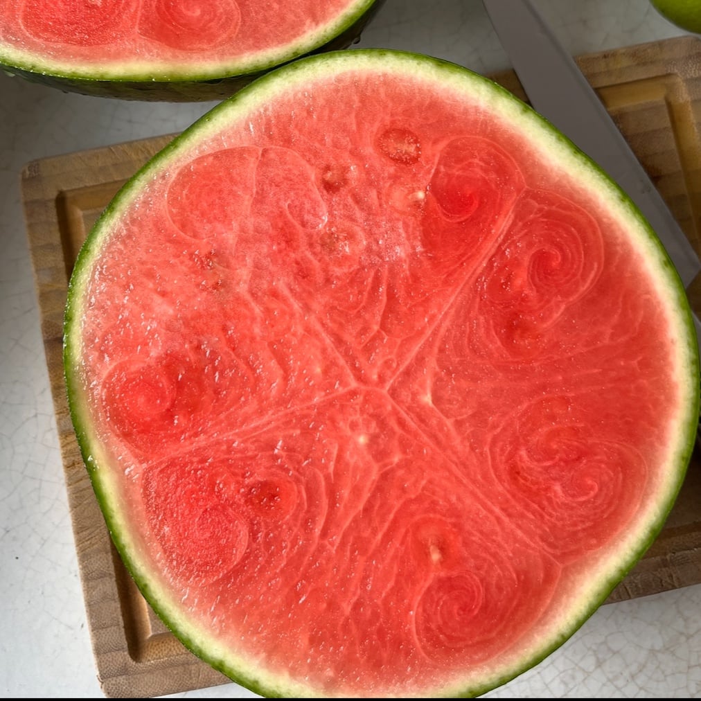 A halved watermelon with an unusual swirling pattern in the red flesh sits on a wooden cutting board, accompanied by a few fresh watermelon slices and a knife partially visible nearby.