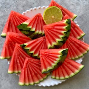A plate of neatly arranged, fan-cut watermelon slices with a half lime on a light-colored surface.