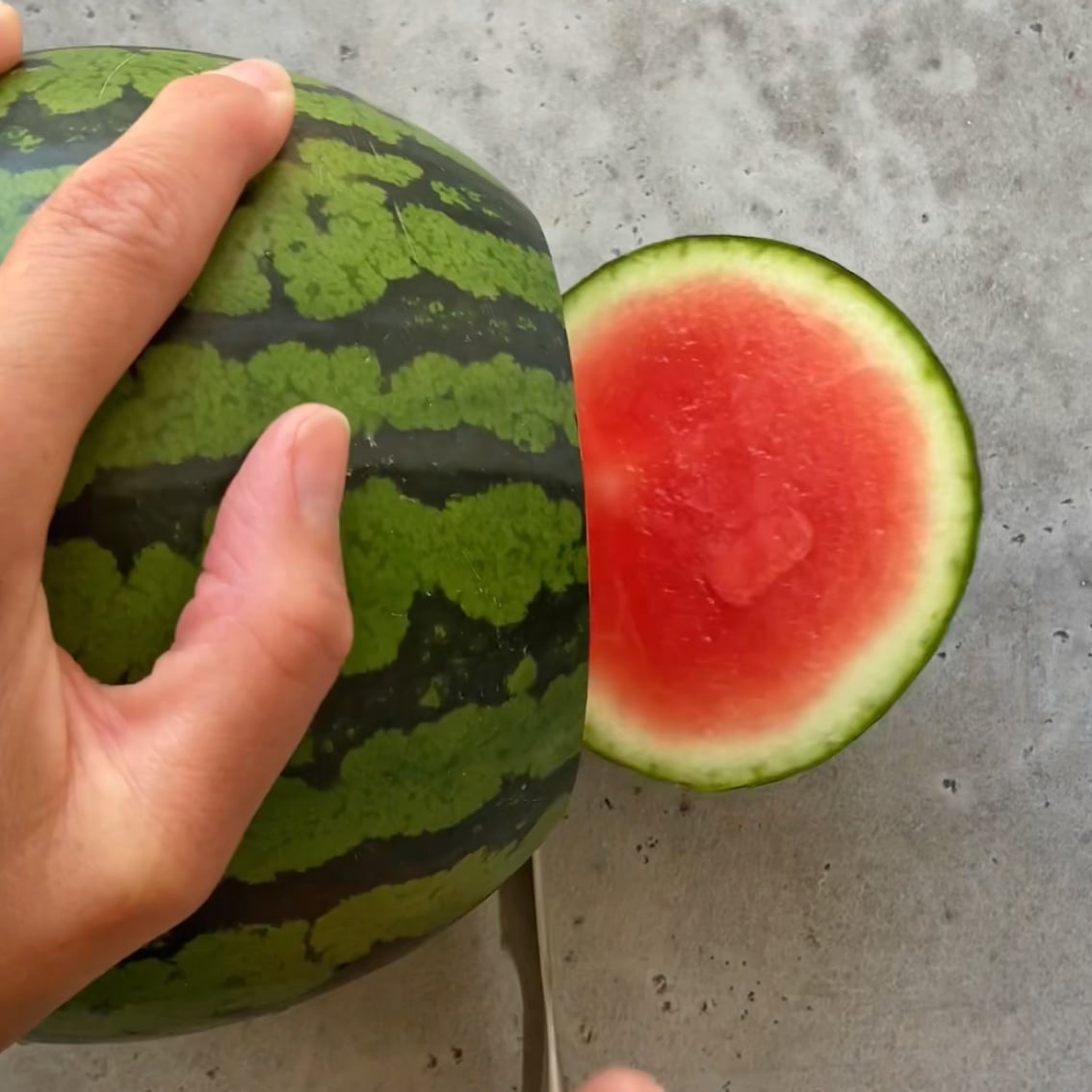 A hand holds a watermelon while a knife slices off a round piece, revealing the red flesh.