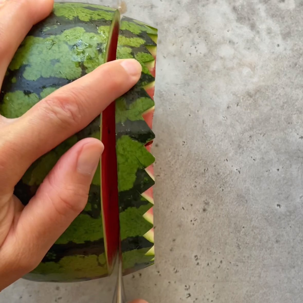 A hand slices a watermelon with a knife, creating a zigzag pattern along the edge.