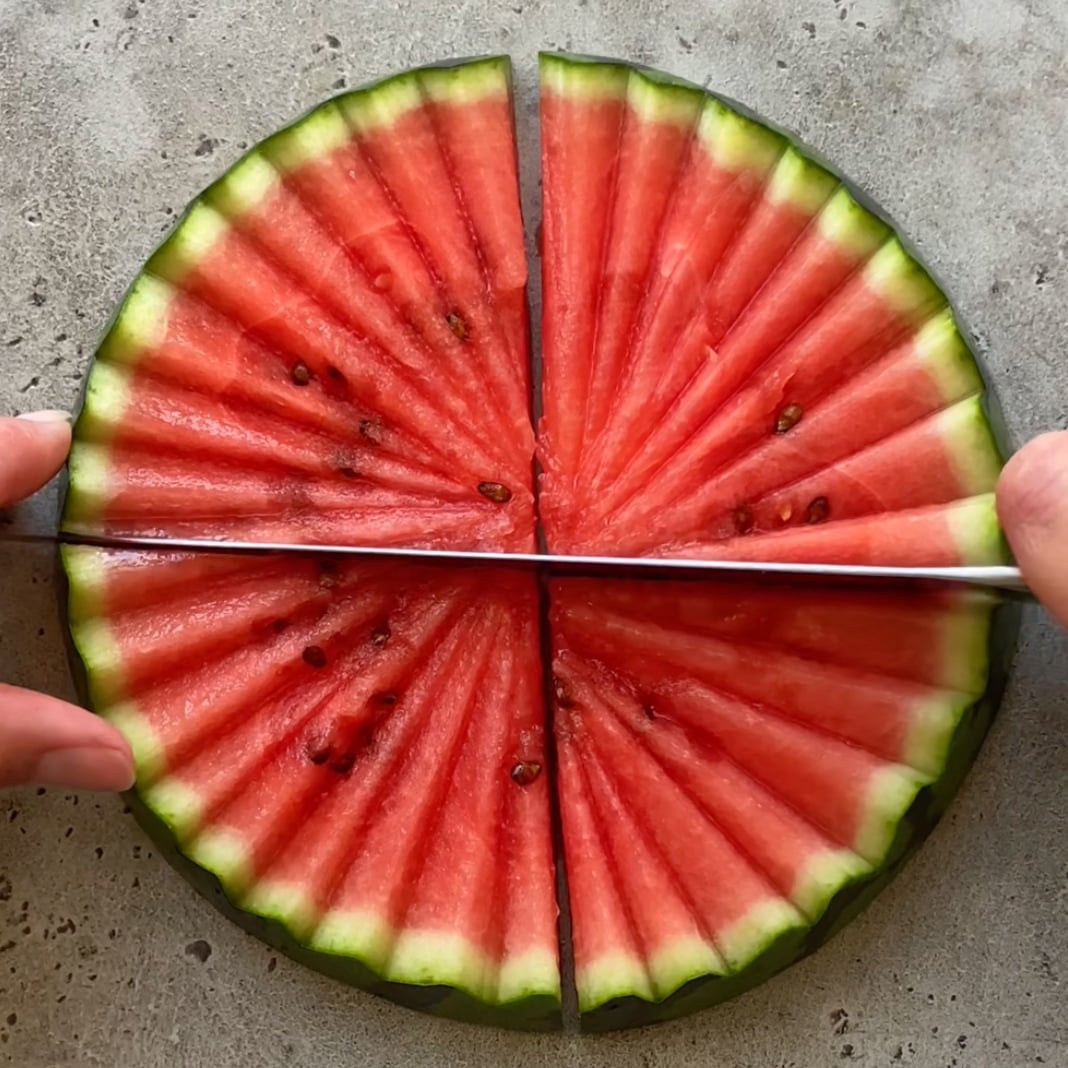 A round slice of watermelon is being cut into quarters, creating perfectly symmetrical Watermelon Slices with vibrant red flesh, scattered seeds, and a crisp green rind resting on a gray surface.