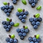 Clusters of blueberries arranged on a gray surface, each with a small basil leaf and some chocolate at the base to resemble miniature grape bunches.