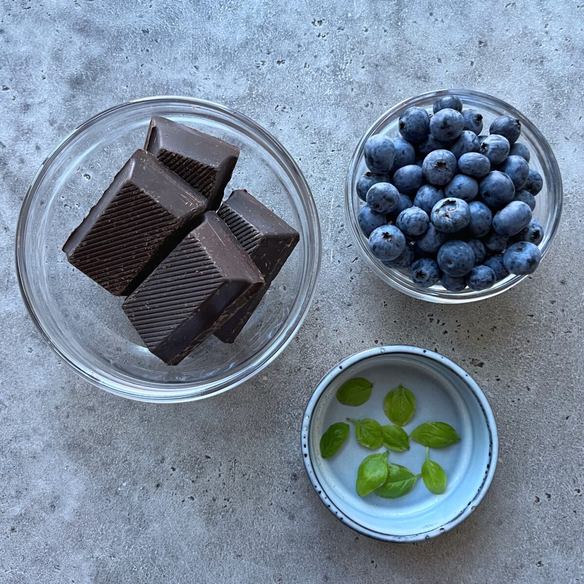 Three glass bowls contain dark chocolate blueberries, fresh blueberries, and green basil leaves on a gray textured surface.
