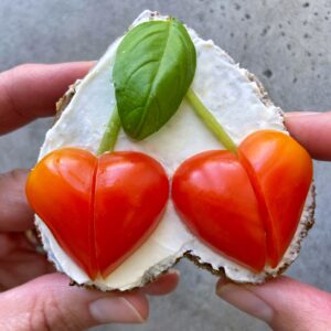 A slice of bread with cream cheese, topped with two heart-shaped tomato halves and a basil leaf, held by two hands.