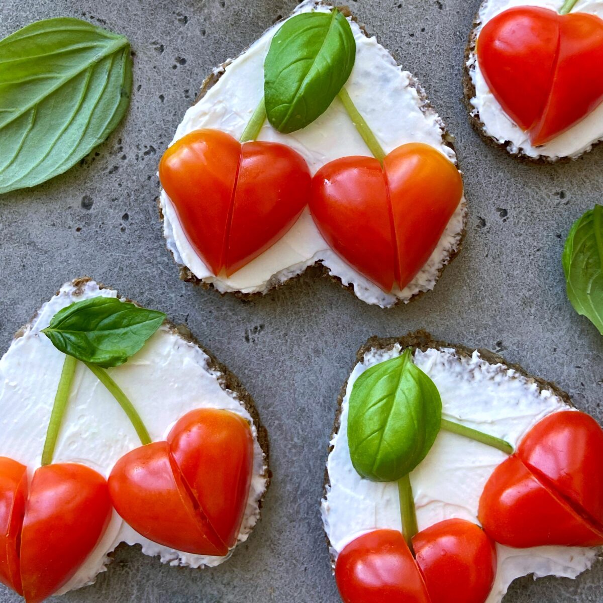 Slices of bread with cream cheese, topped with heart-shaped cherry tomatoes arranged like cherries, and garnished with fresh basil leaves.
