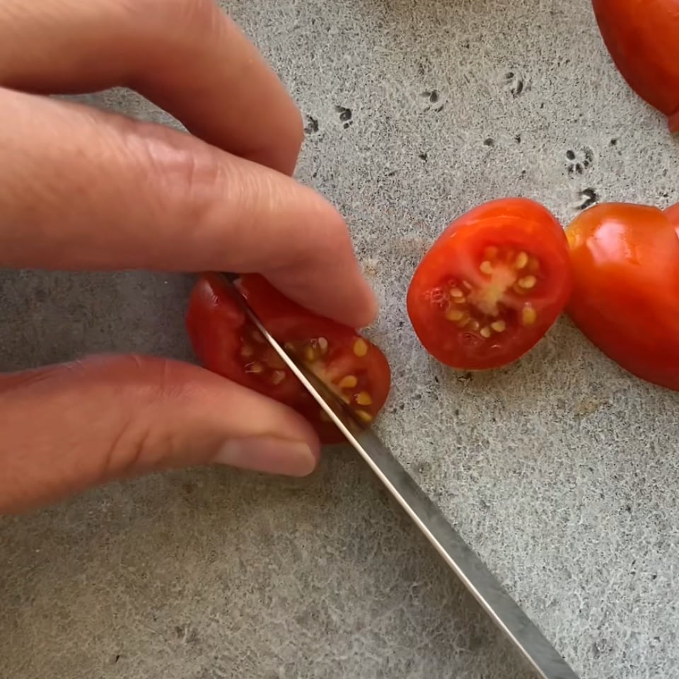 A knife slices two angled grape tomato slices in half.