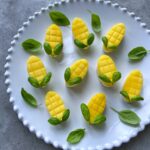 A white plate holds nine Feta & Mango Appetizers, each piece of mango cut to resemble small corn on the cobs with crosshatch patterns.