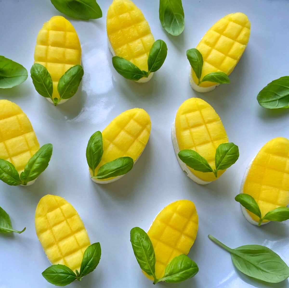 Sliced yellow mango pieces with corn patterns are arranged on a white plate, each piece decorated with two green basil leaves.