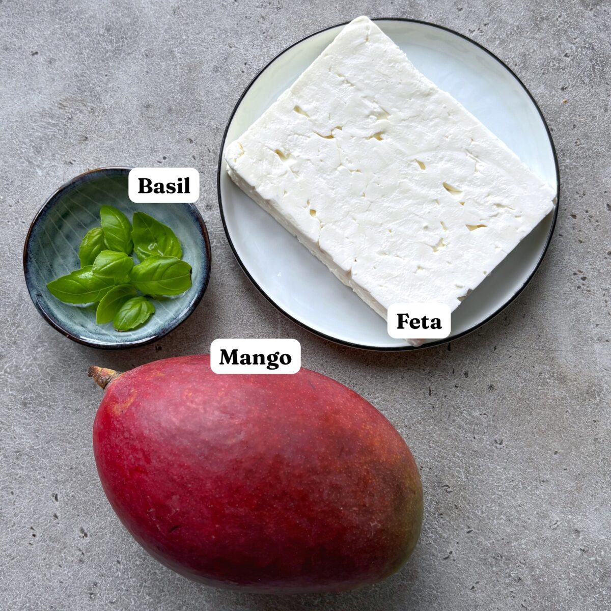 A whole mango, a small bowl of fresh basil, and a block of feta cheese.