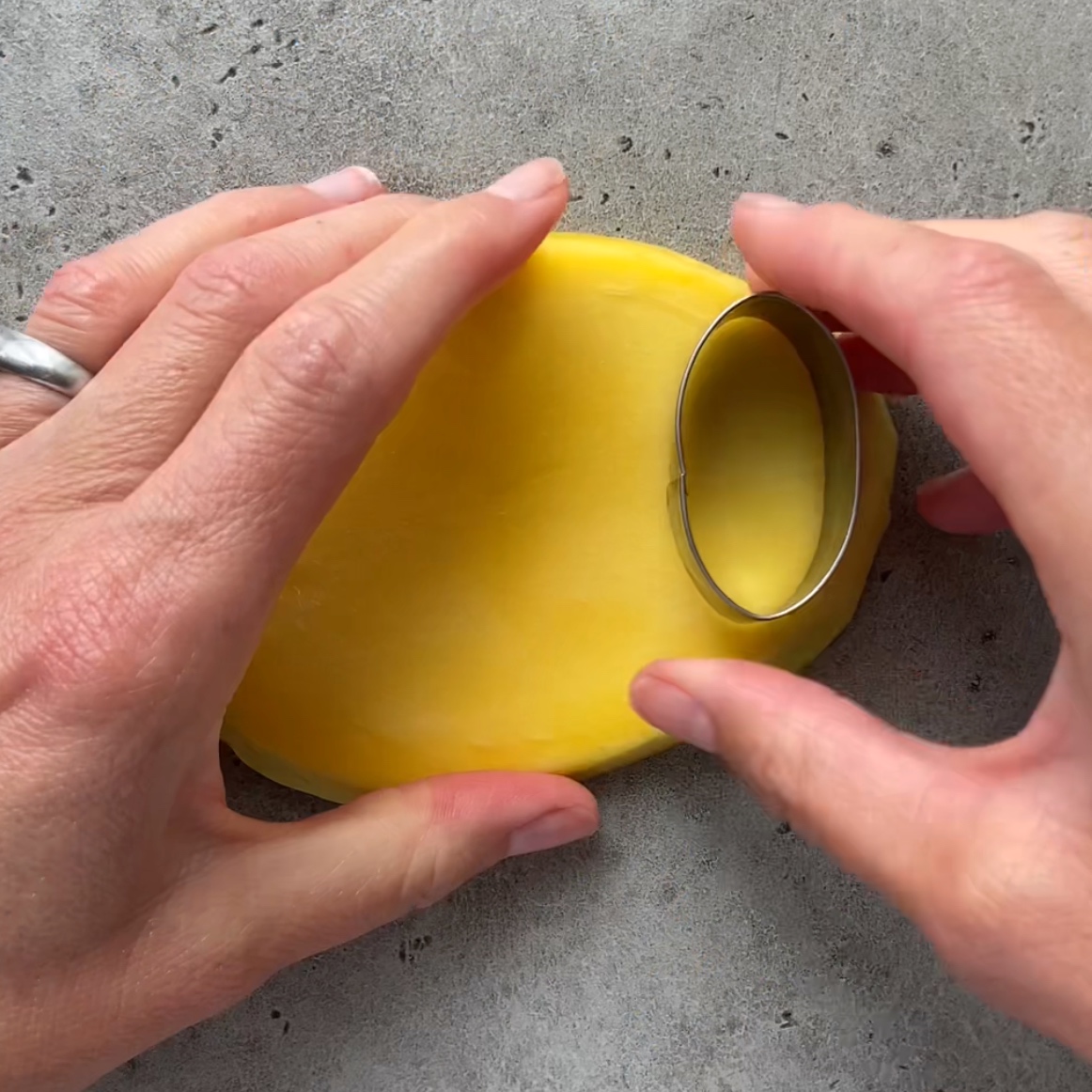 Two hands use an oval shaped cookie cutter to cut mango on a grey surface.