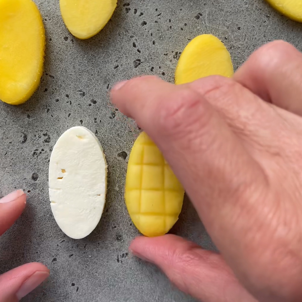 A hand holds an oval mango slice scored with a grid pattern next to an oval slice of feta.