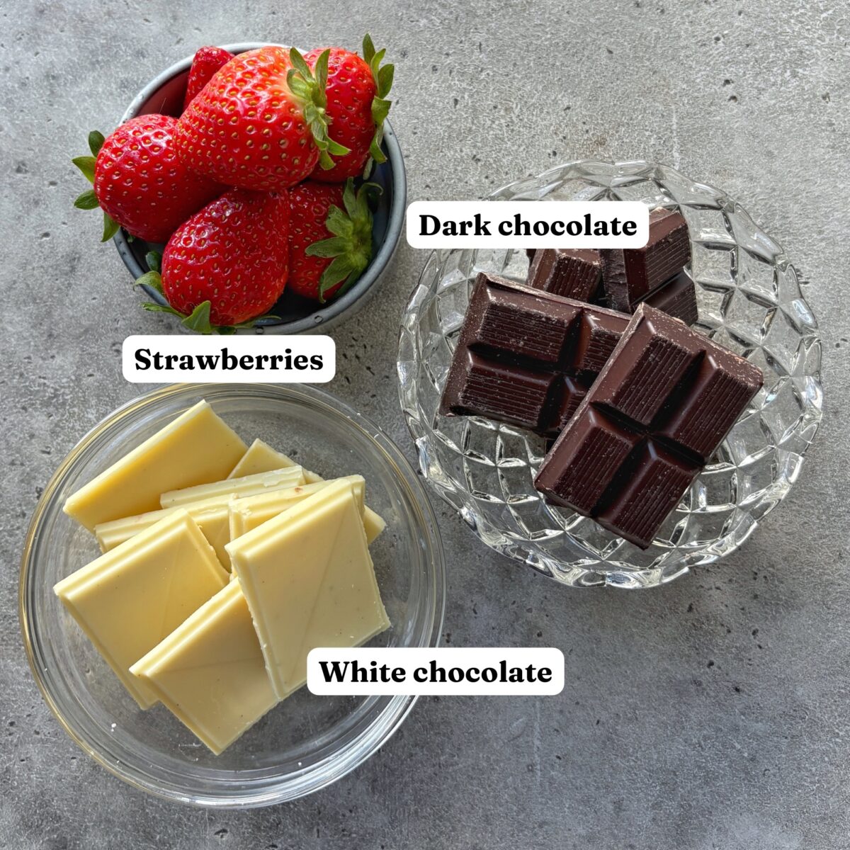 Three glass bowls containing fresh strawberries, squares of dark chocolate, and squares of white chocolate on a gray surface. Each item is labeled.